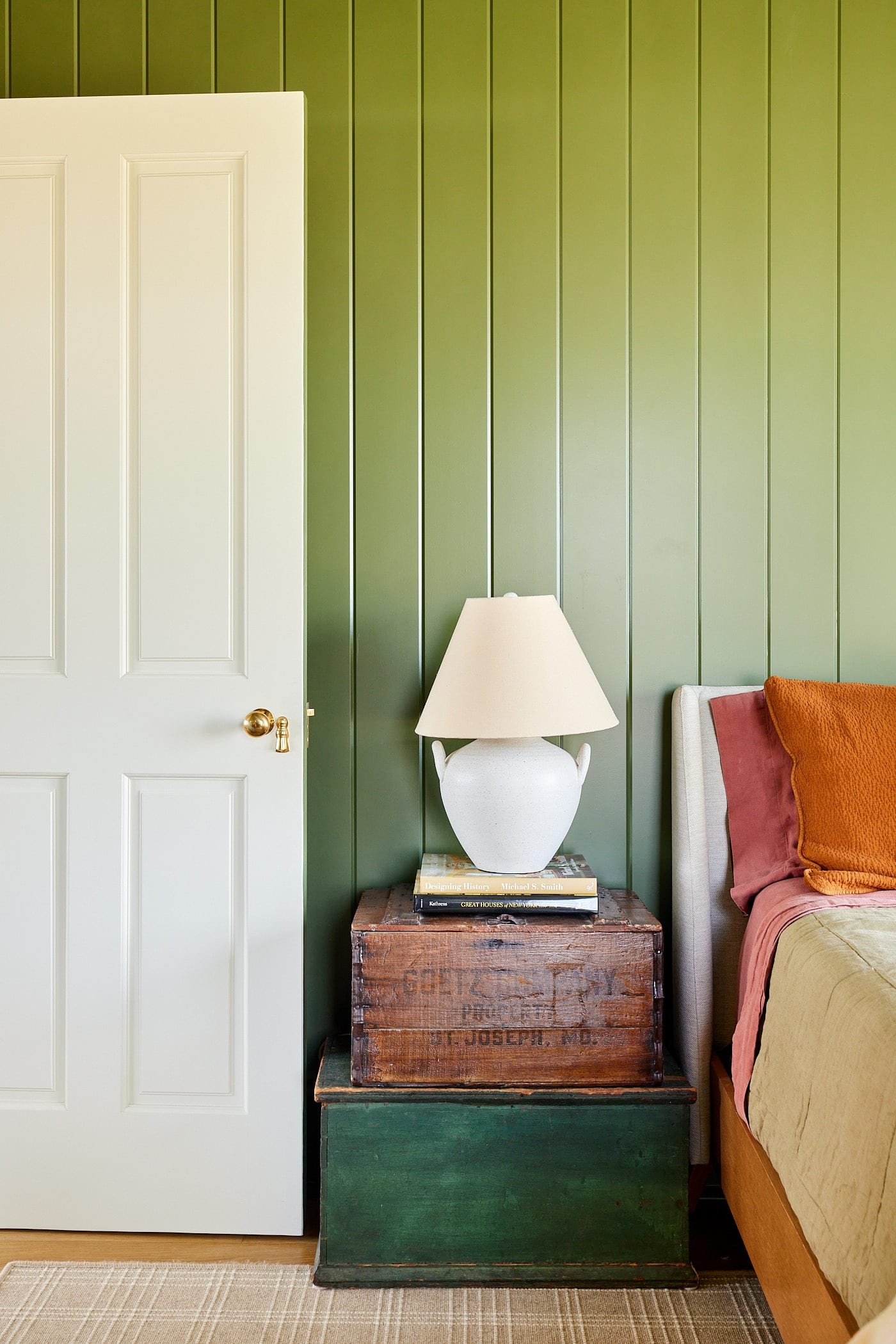 Green walls with tongue and groove with bed to the side and two antique boxes acting as an end table with white lamp on top.
