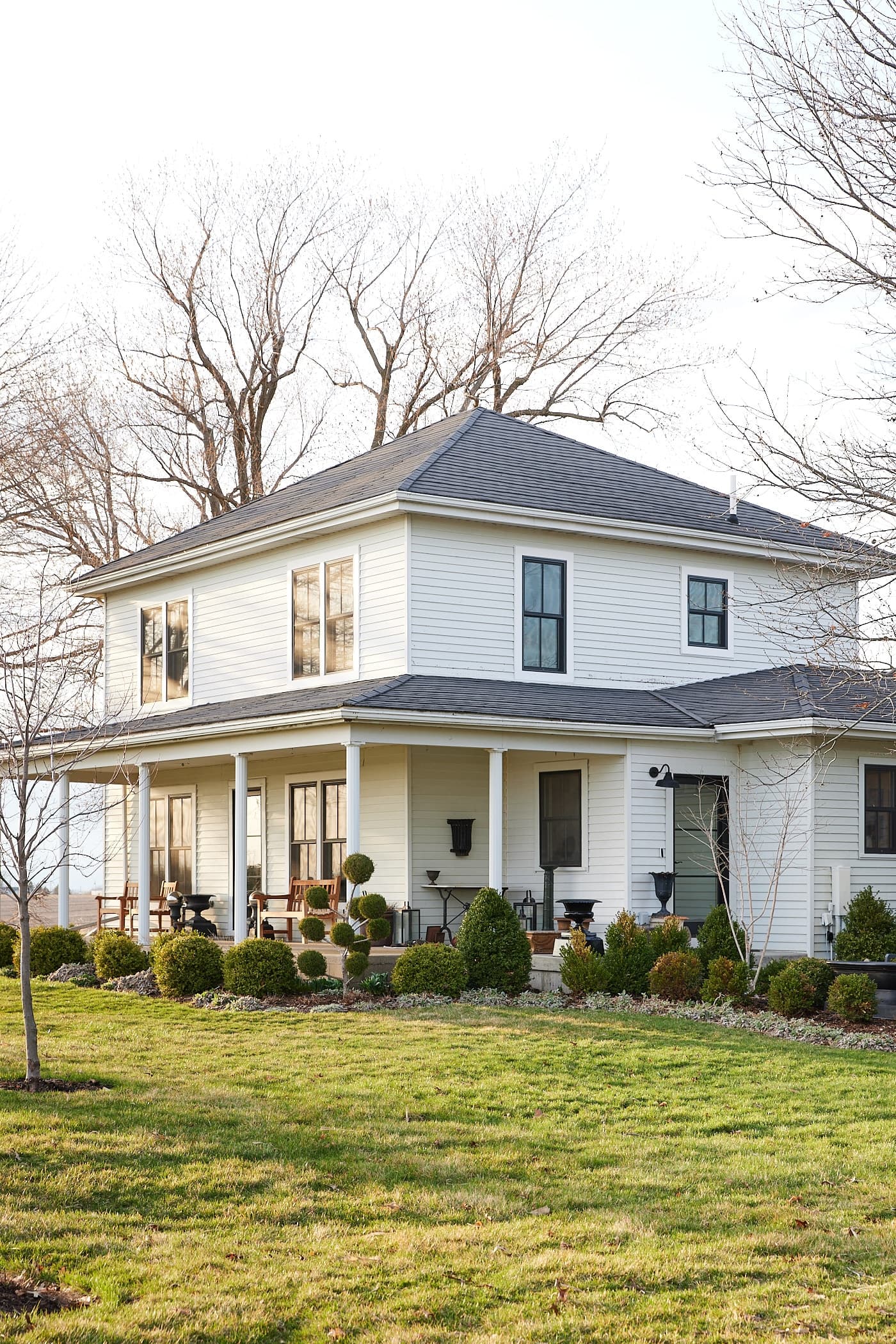 White farmhouse with black windows and white trim with green grass and lots of landscaping out front with no leaves on the trees.