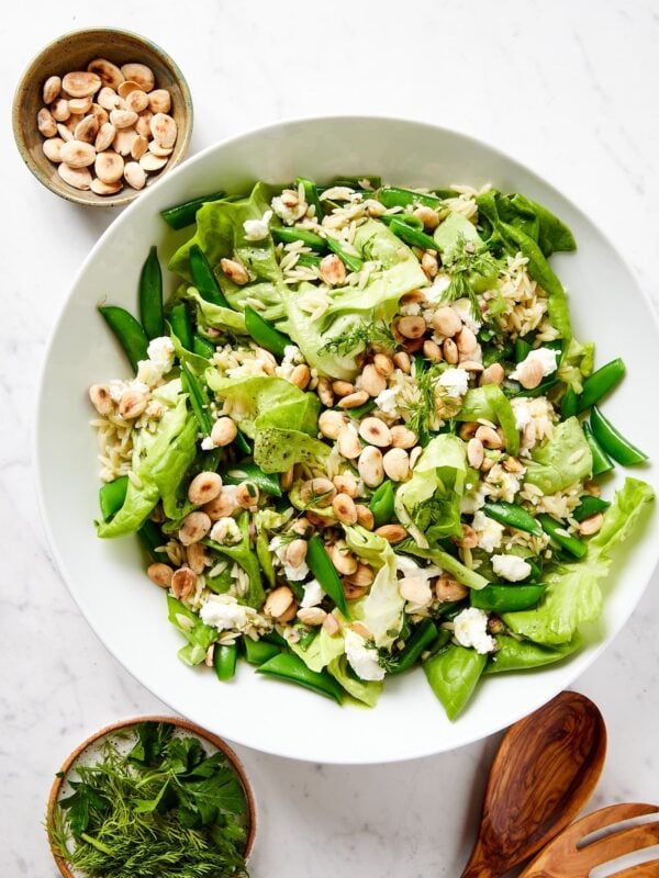 White salad bowl filled with butter lettuce, snap peas, orzo, and toasted Marcona almonds all on white marble surface.