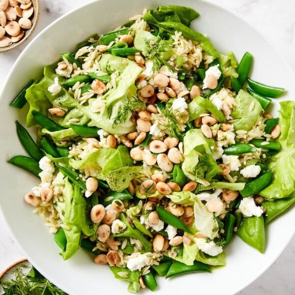 White salad bowl filled with spring orzo salad with toasted Marcona almonds and goat cheese sprinkled on top all on white marble surface.