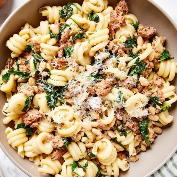 Brown bowl on marble surface with striped cloth filled with cooked pasta mixed with spinach and sausage mixture.