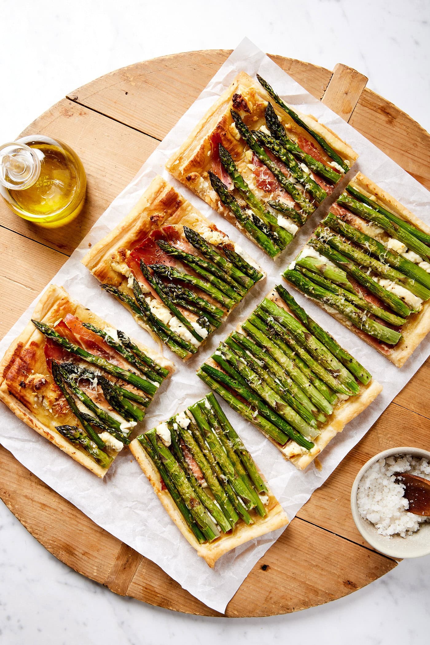 Squares of tart on a piece of parchment paper with prosciutto underneath asparagus and sprinkled with cheese.