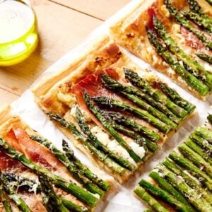 Pieces of asparagus tart sitting on a wooden board with parchment underneath with baked pieces of asparagus and puff pastry.
