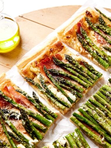 Pieces of asparagus tart sitting on a wooden board with parchment underneath with baked pieces of asparagus and puff pastry.