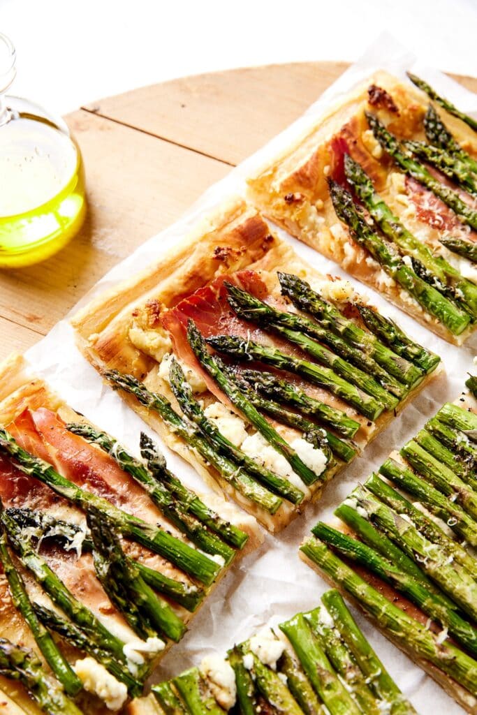 Pieces of asparagus tart sitting on a wooden board with parchment underneath with baked pieces of asparagus and puff pastry.