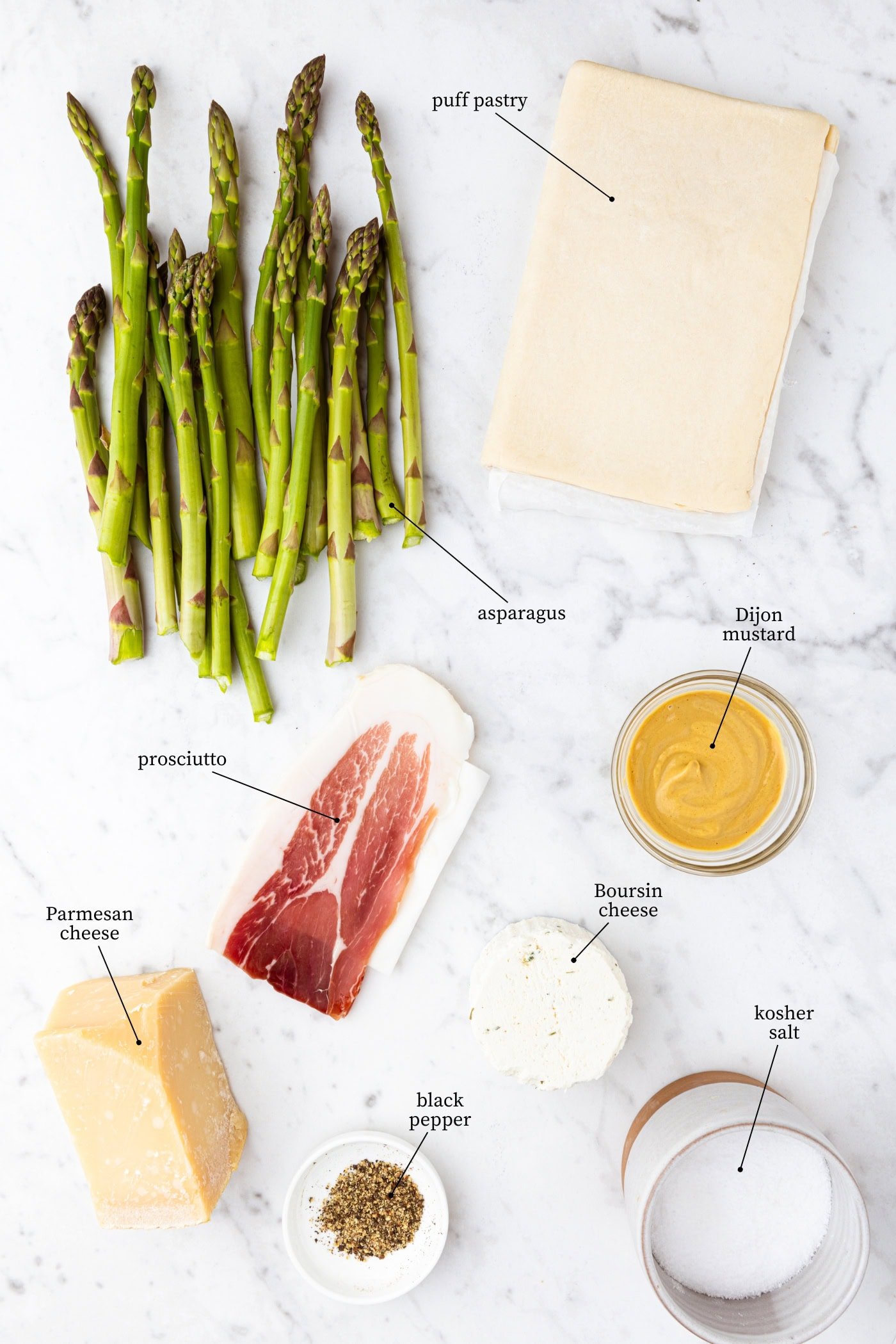White marble surface filled with all ingredients needed to make a tart including asparagus, prosciutto, cheese, puff pastry, and more.
