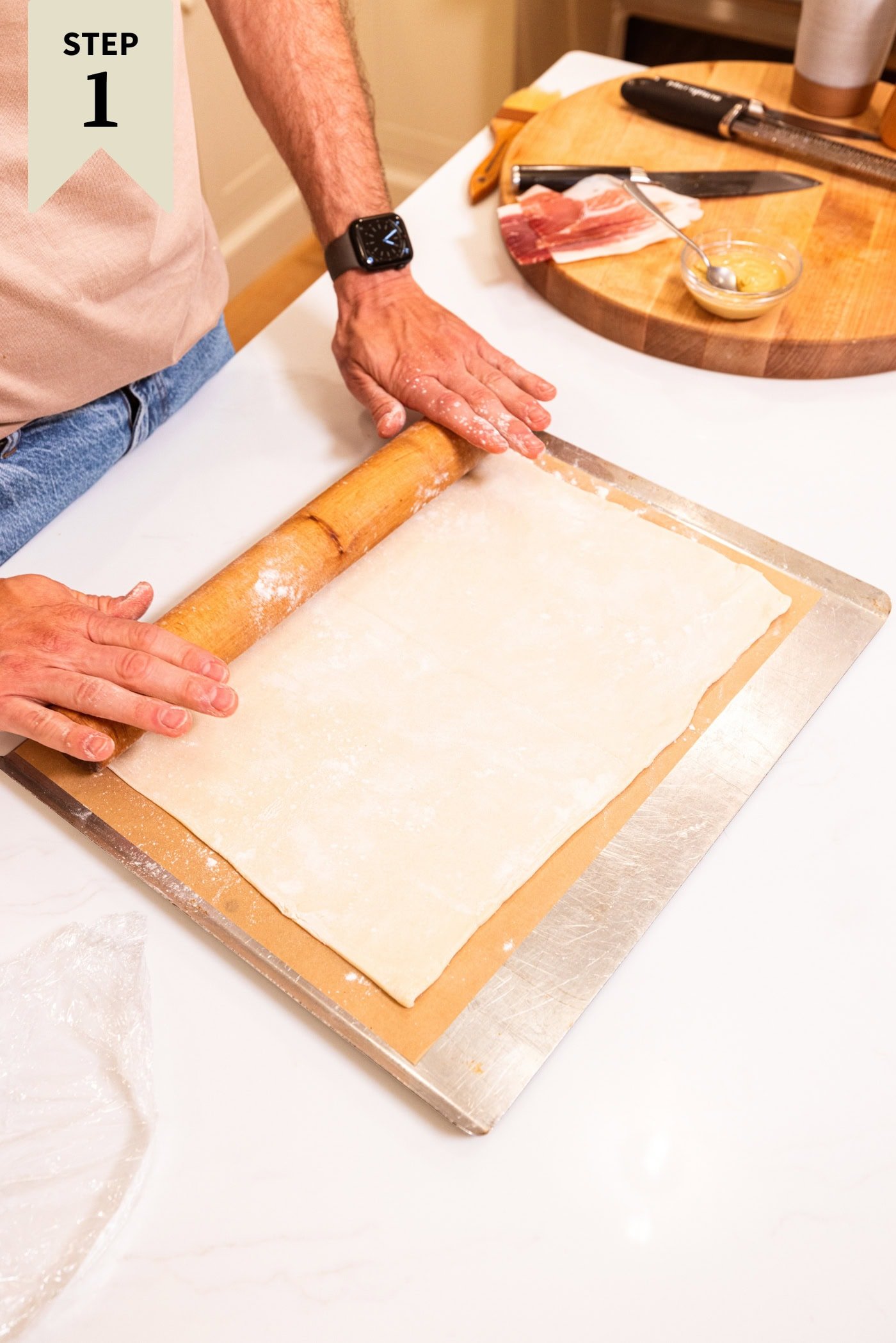 Puff pastry being rolled out on a sheet pan with a rolling pin to form an asparagus tart.