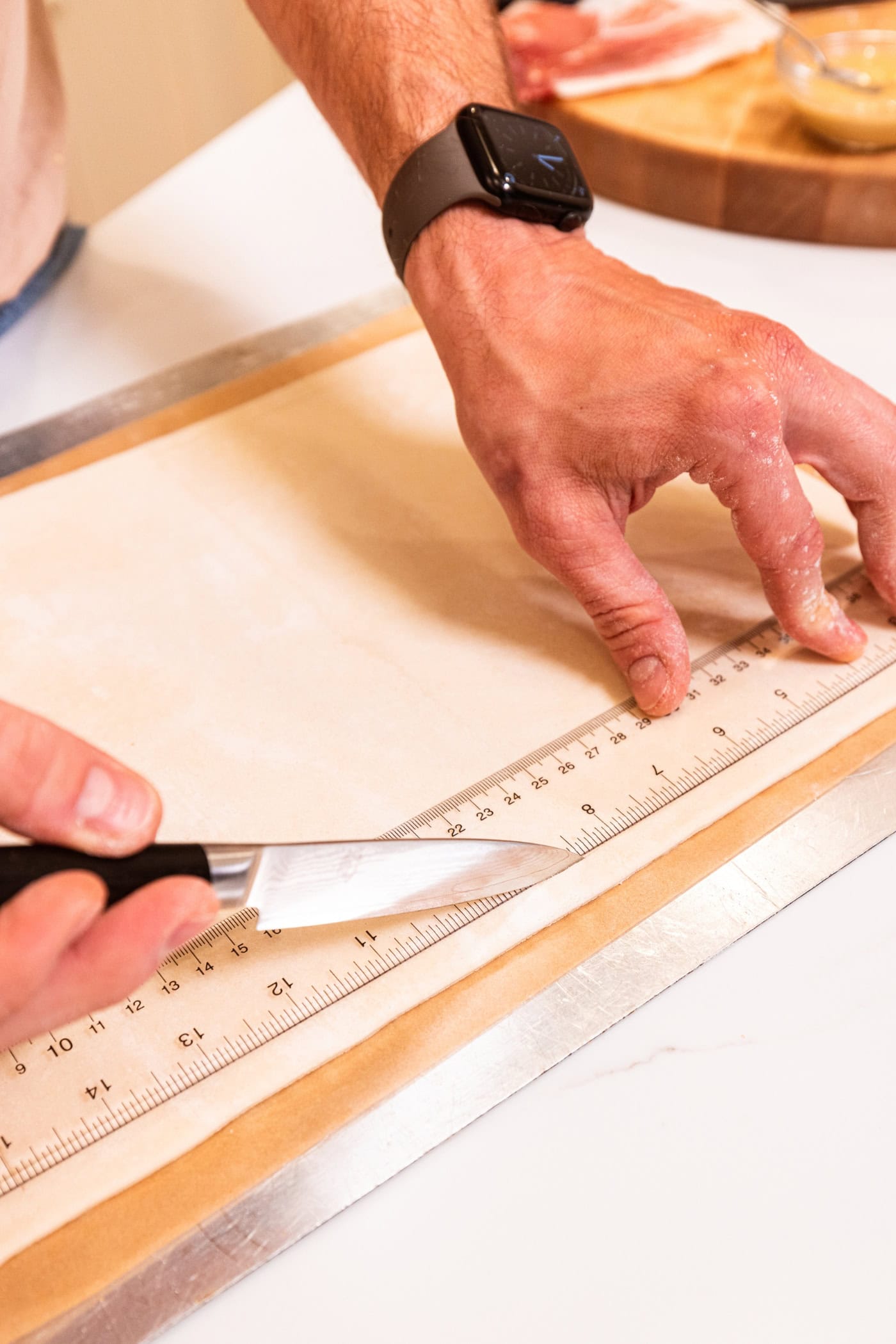 Hand holding knife and a ruler slicing into puff pastry along the side of the ruler.
