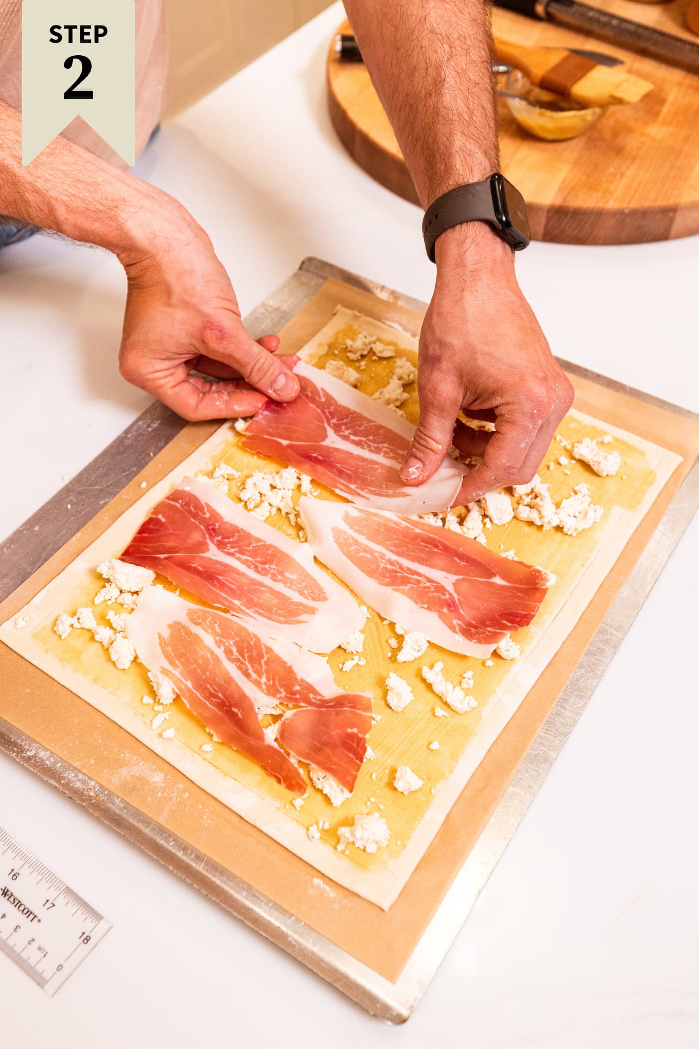 Hands placing pieces of prosciutto on top of cheese and Dijon mustard on puff pastry to create an asparagus tart.