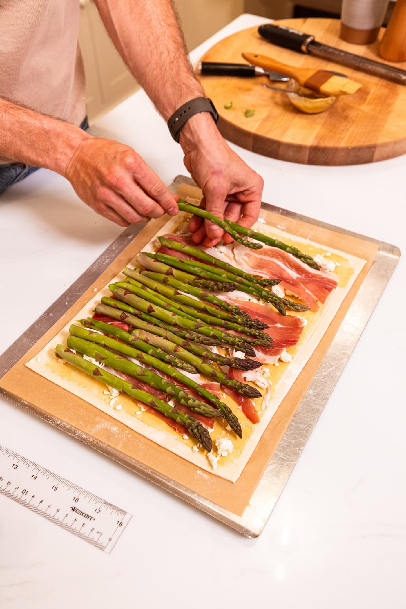 Two hands placing pieces of asparagus on top of prosciutto and cheese and Dijon mustard on puff pastry for a tart.