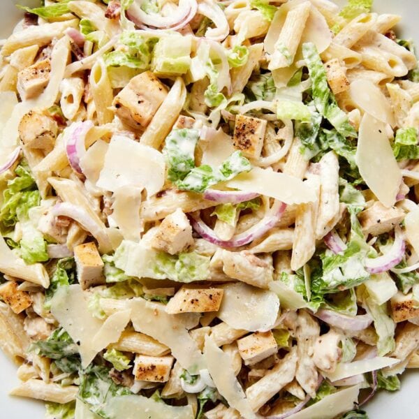 White bowl filled with chicken Caesar pasta salad with shards of Parmesan cheese, lettuce, red onion, and tossed in Caesar dressing.