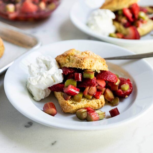 Strawberry and rhubarb shortcakes with whipped cream on two bowls with golden biscuits and macerated fruit in a bowl in the background on a marble surface.