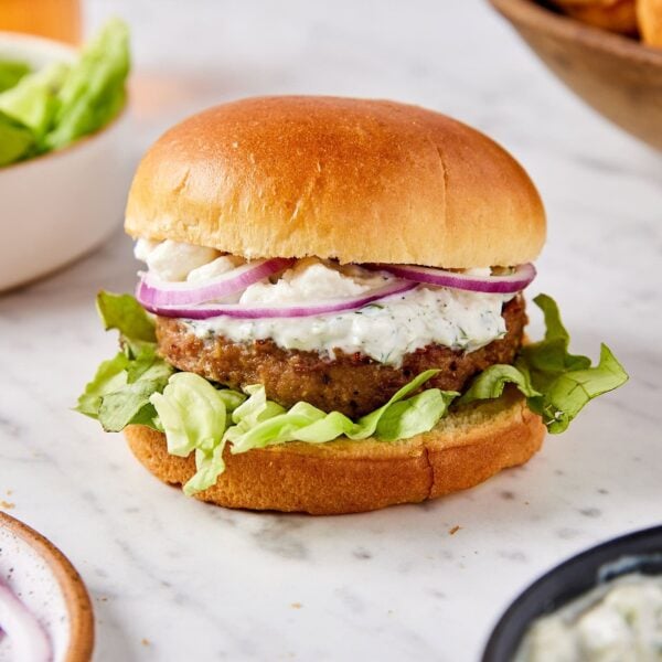 White marble surface with Greek turkey burger in bun with lettuce, feta cheese, red onions, and tzatziki sauce.