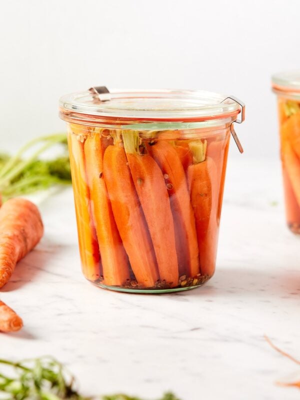 Glass jar filled with quick pickled carrots sitting on marble surface with carrots to the side.
