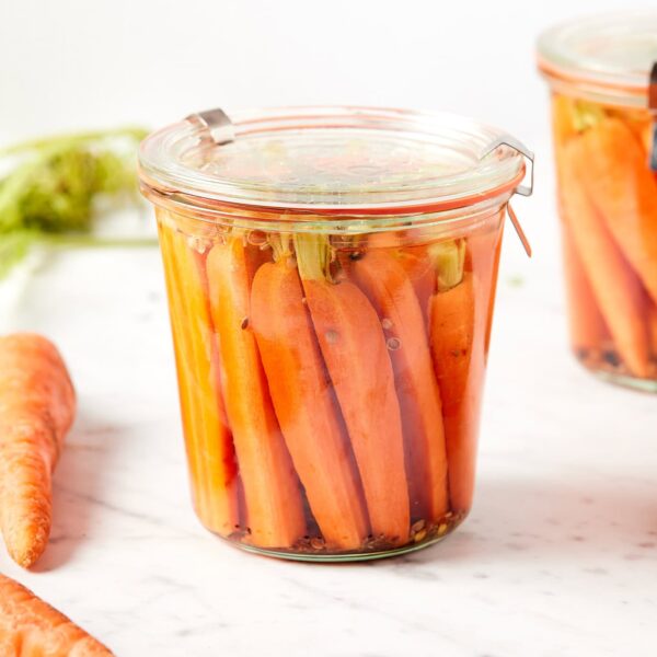 White marble surface with jar of quick pickled carrots with more carrots all around including another jar.