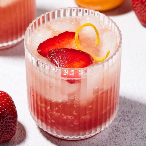 Glass filled with red color cocktail with slices of strawberry on top along with a sliver of orange peel.