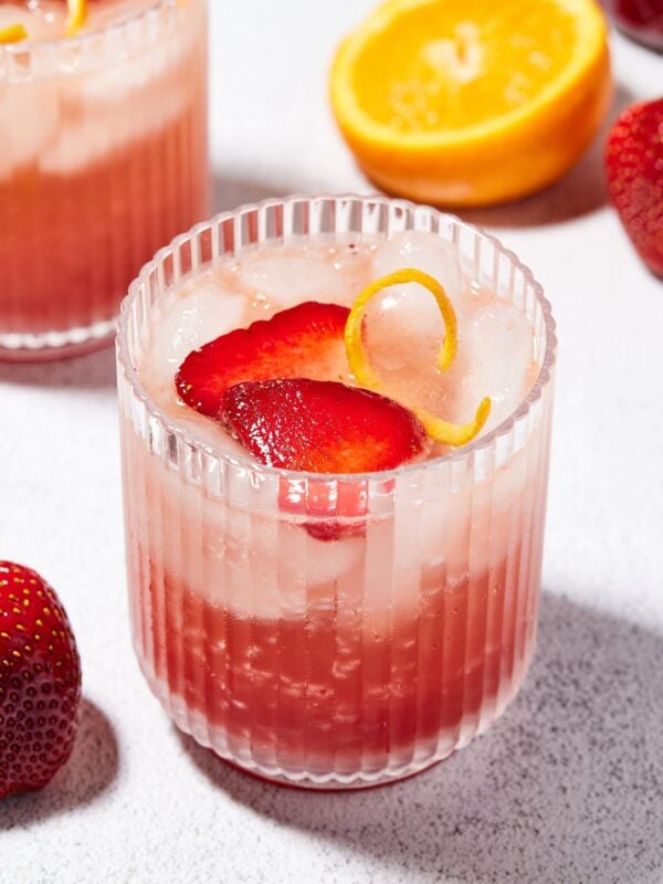 Glass filled with strawberry gin spritz with orange peel and pieces of strawberry with another glass in the background.