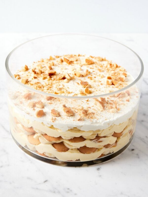 Glass trifle dish filled with banana pudding recipe with crumbled vanilla wafer cookies on top on white marble surface.