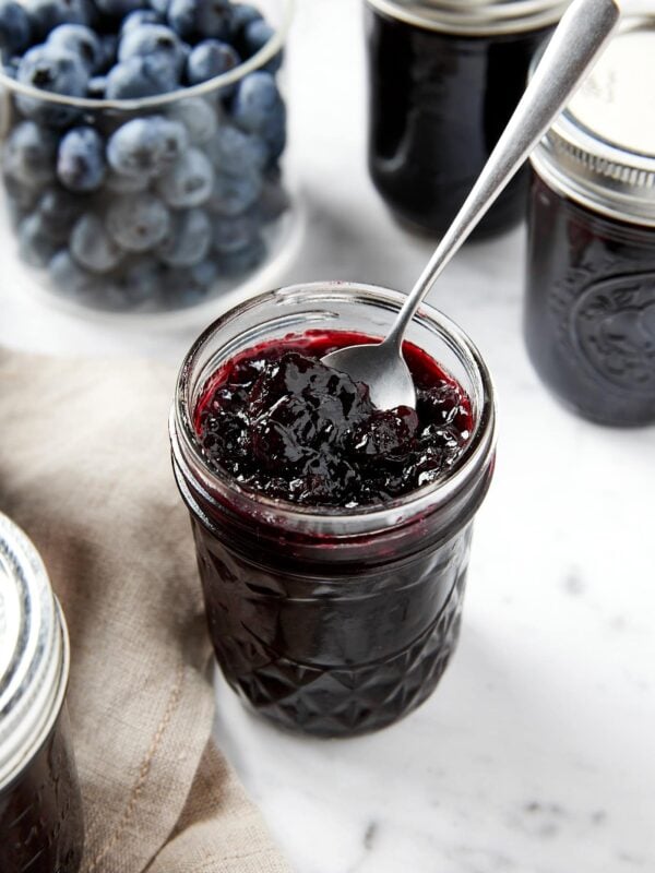 Small jam jar filled with blueberry jam with more raw blueberries in background with spoon resting in jar.