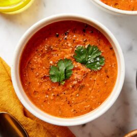 White bowl filled with gazpacho soup recipe with fresh parsley leaves on top on white surface.
