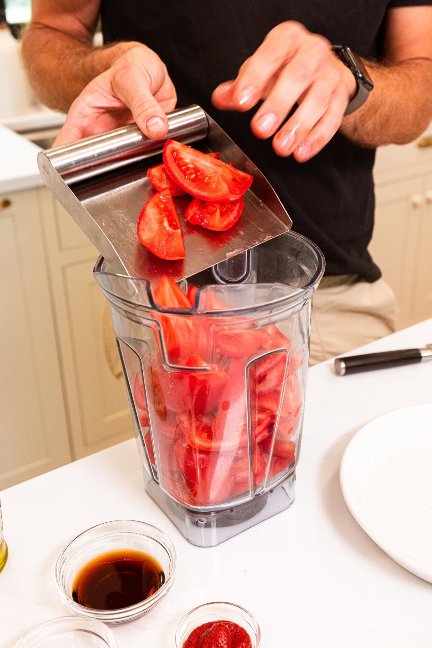 Hand using metal scraper to push chopped tomatoes into a large canister blender.