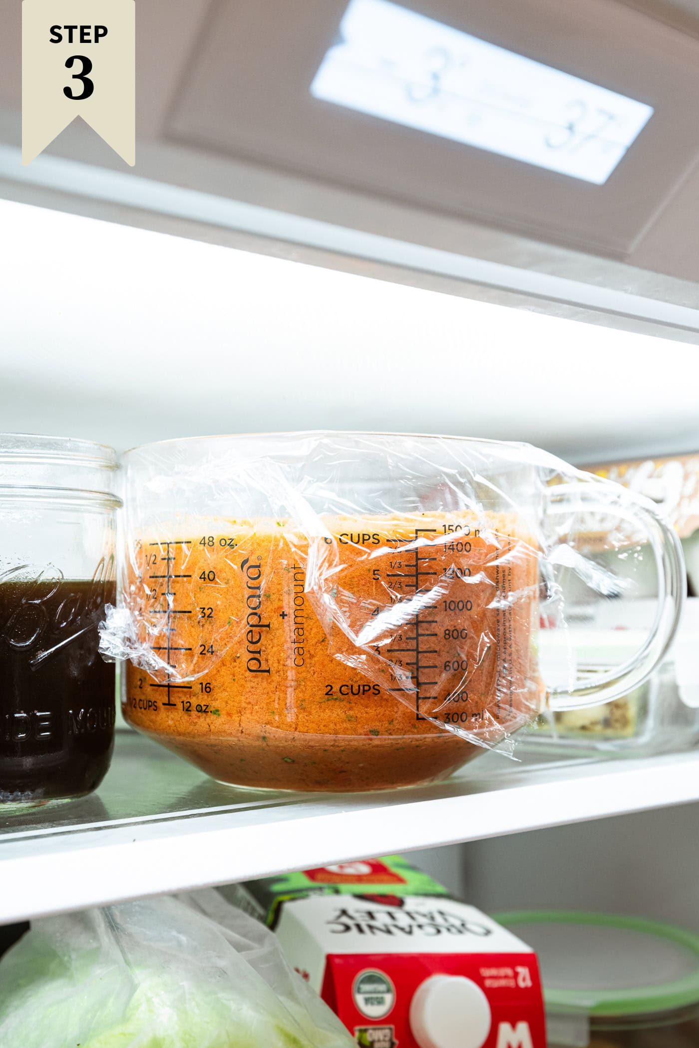 Measuring cup with plastic on top sitting in the fridge with red tomato soup in it to chill.