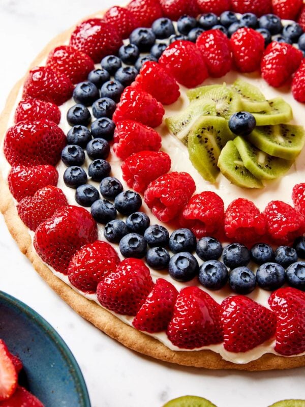 Round fruit pizza topped with strawberries, blueberries, raspberries, and more fruit on white marble surface.