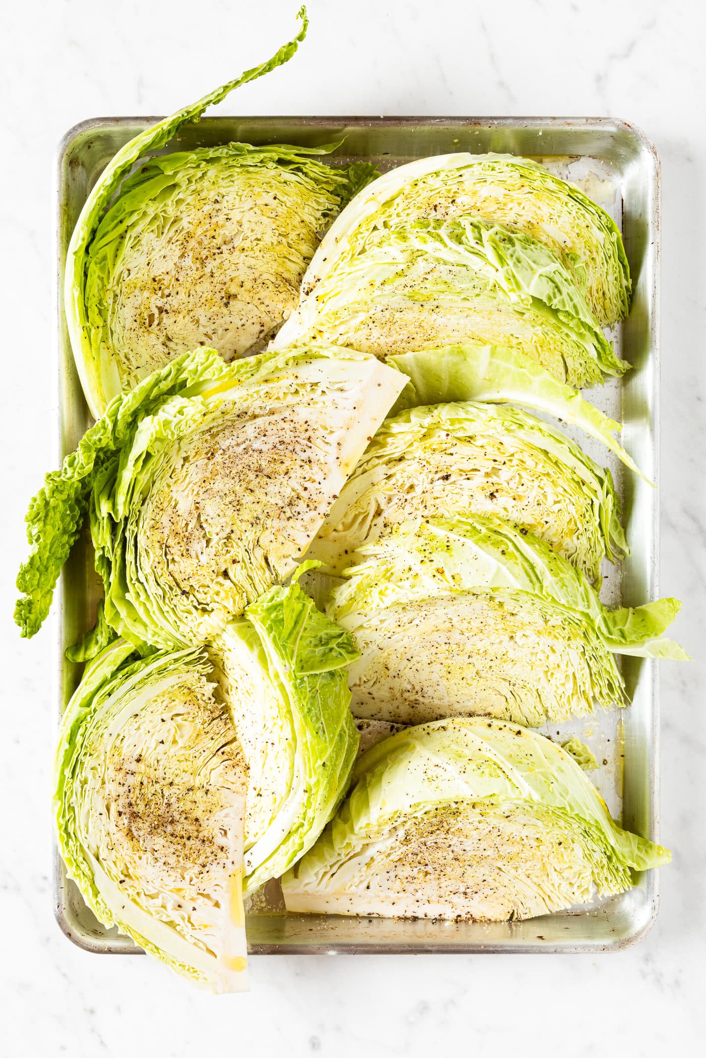 Metal sheet pan filled with wedges of cabbage with black pepper, salt, and oil on top.