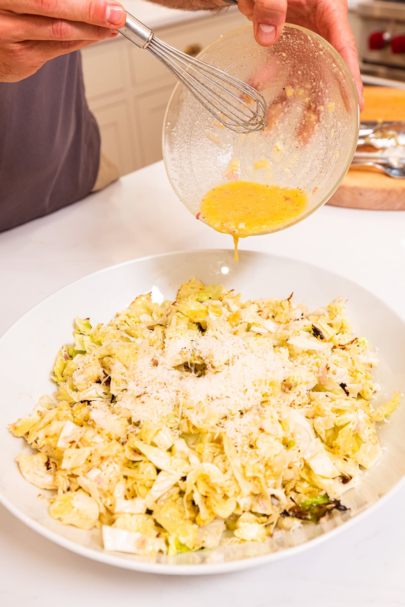 Hand pouring Parmesan dressing over the top of chopped cabbage from a glass bowl.
