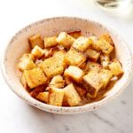 White bowl filled with homemade croutons with crispy exterior on white marble surface.