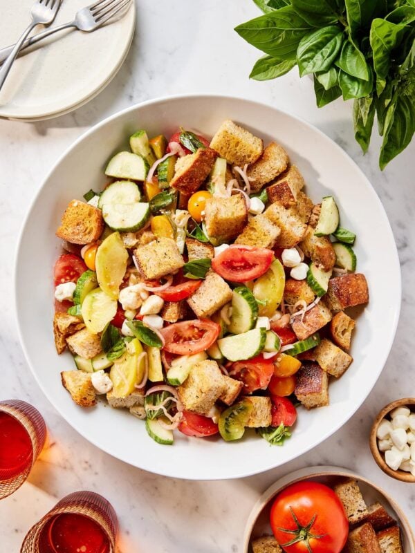 Large white bowl filled with panzanella salad all on white surface with plates and fresh basil around.