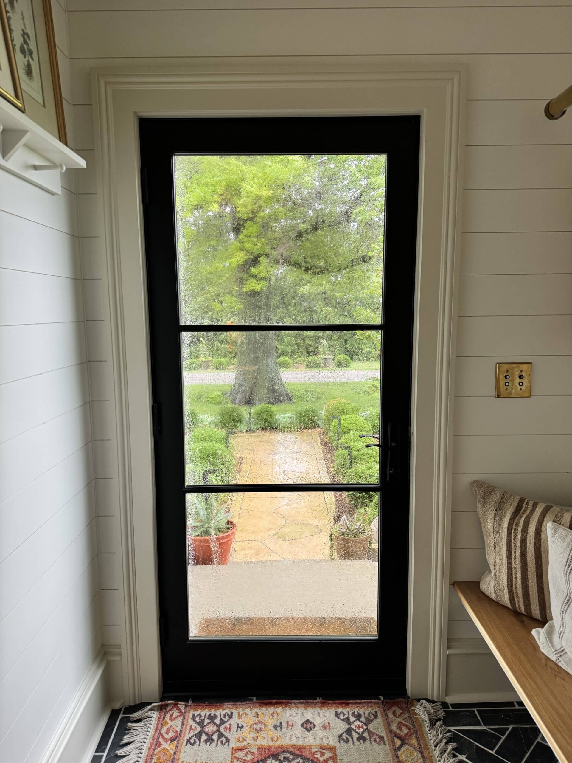 Black front door with rain dripping down the glass with bench with pillows on it inside.