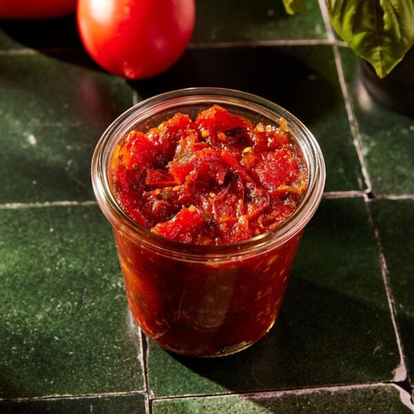 Glass container filled with tomato jam after cooking on the stove sitting on green tile surface.