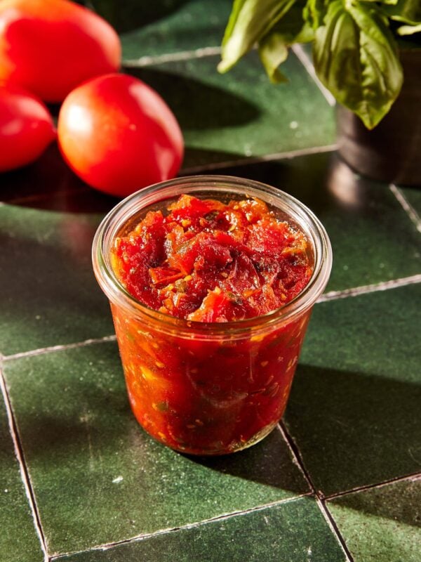 Green tile surface with jar of tomato jam and more tomatoes and basil in background.