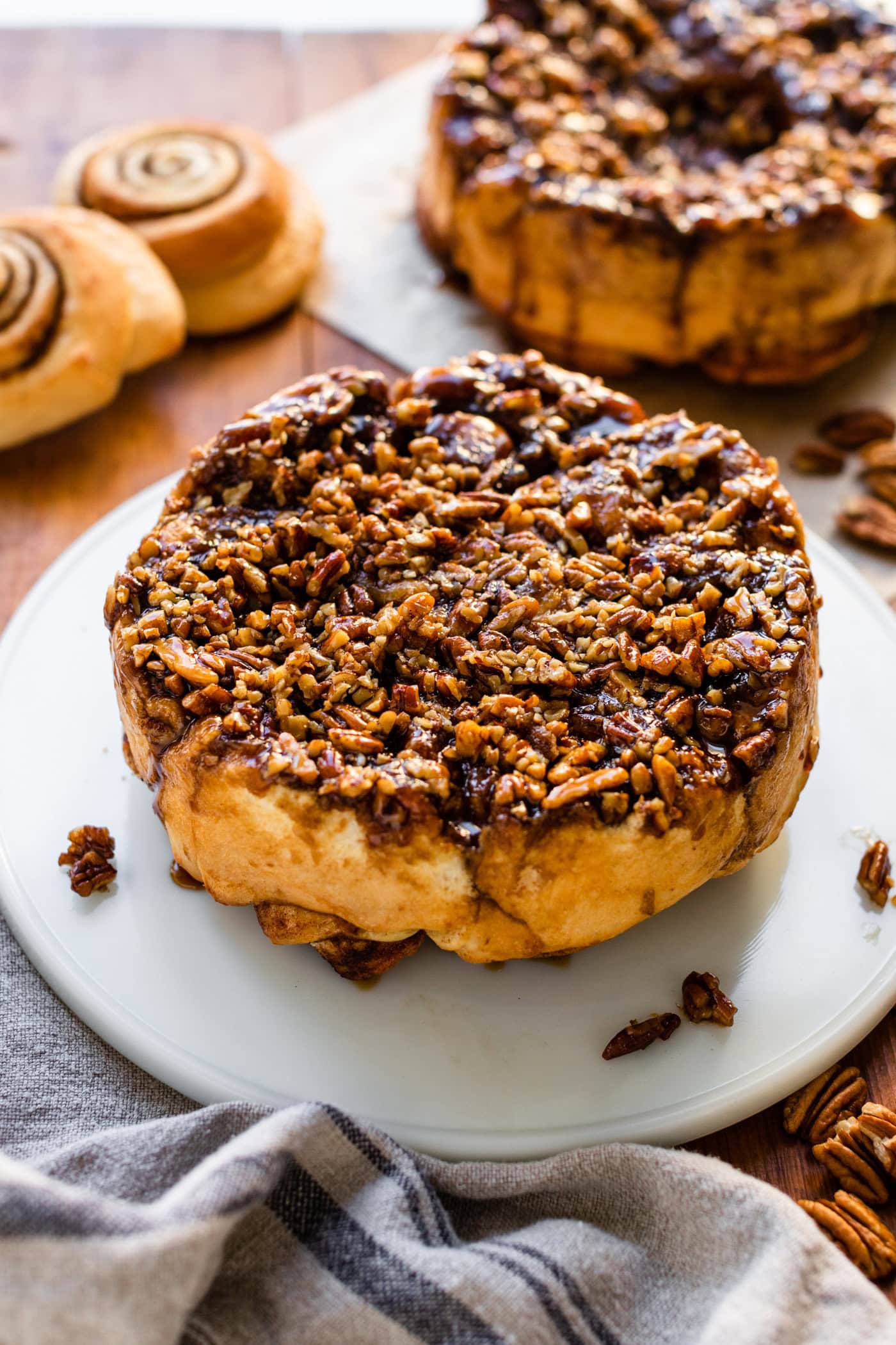 Circle of sticky buns recipe sitting on a white serving platter with nuts sprinkled around with unfrosted cinnamon rolls in background.