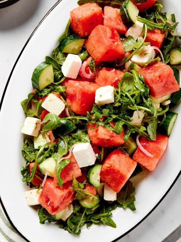 White serving platter with cubes of watermelon sprinkled on top of arugula, feta, and cucumber tossed in a honey and lime dressing.
