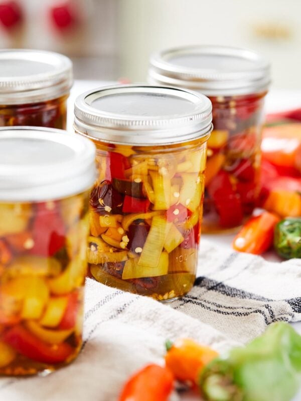 Jars of pickled peppers with lids and rings on top after being canned with more fresh peppers to side.