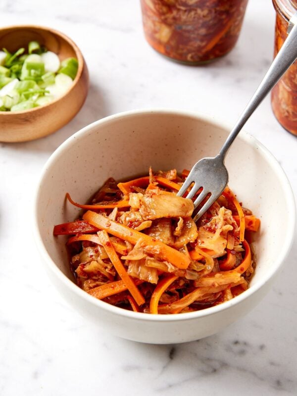 White bowl filled with orange colored kimchi with fork taking a few pieces of cabbage from the bowl all on white marble surface.