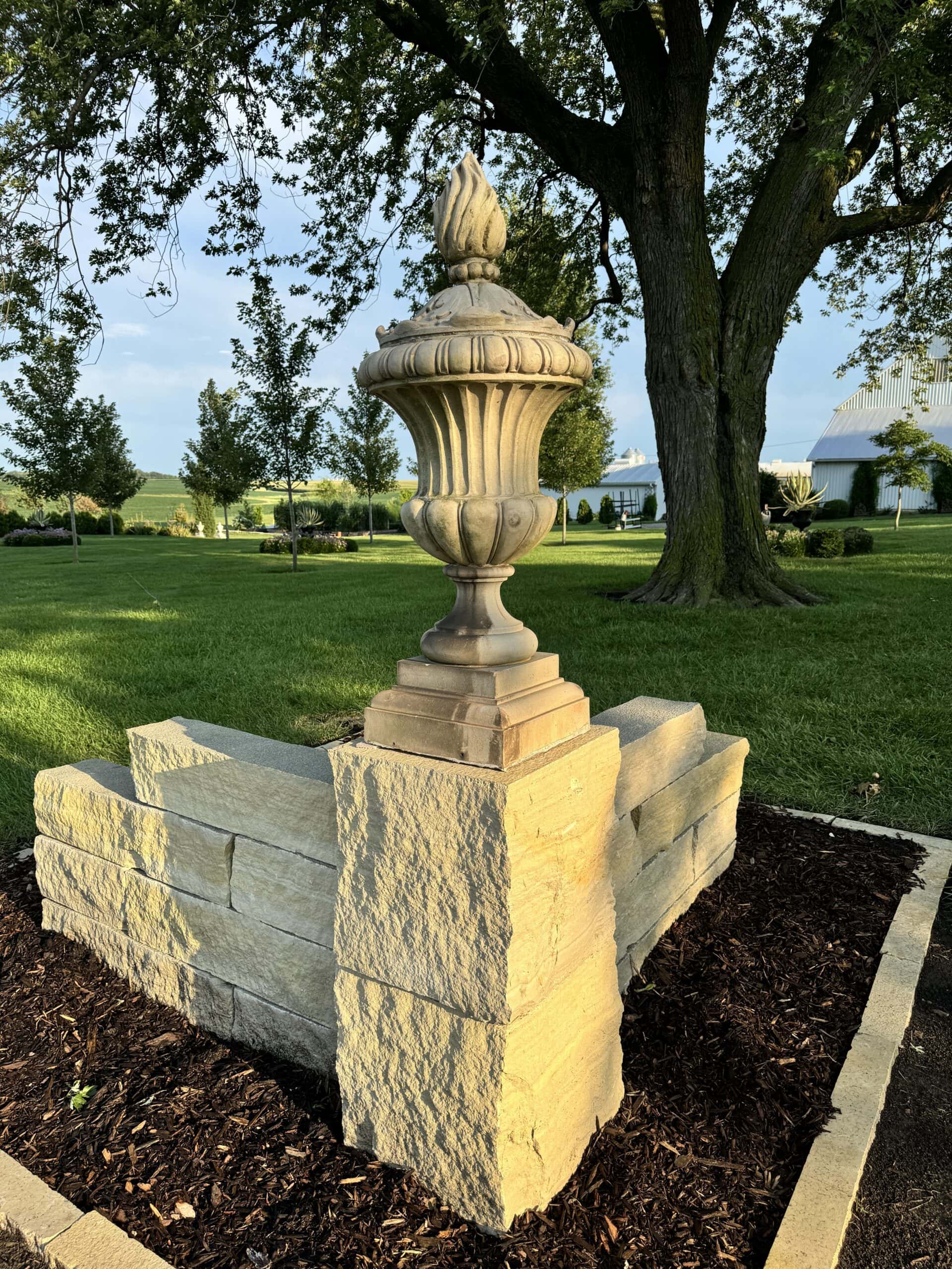 Stone column besides a driveway with a stone urn on top with mulched flowerbed around the base.