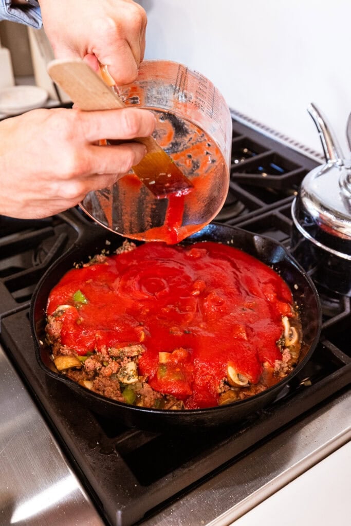 Strained tomatoes get added to a sausage and vegetable mixture on the stove.