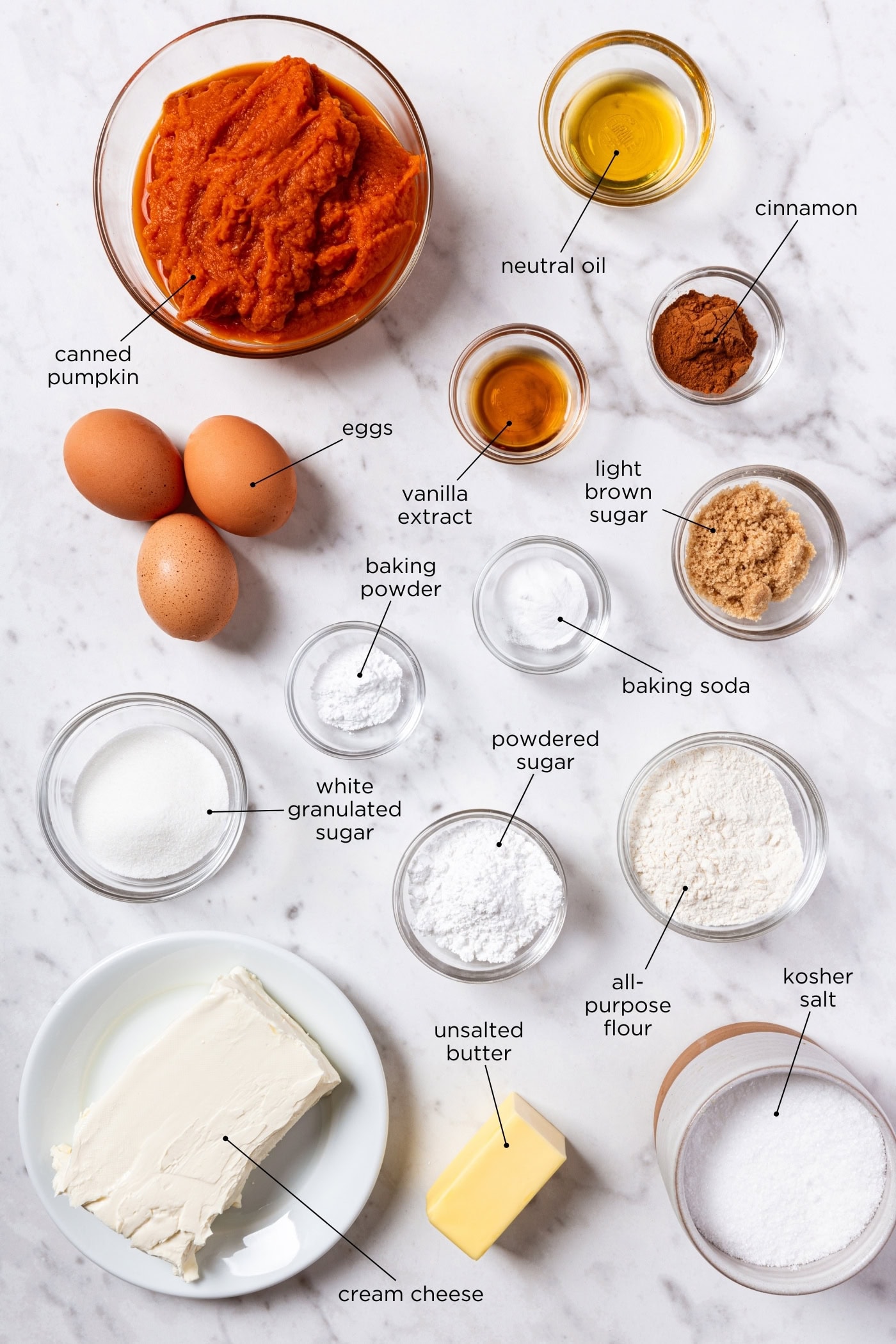 White marble surface with ingredients for pumpkin bars including eggs, spices, flour, sugar, pumpkin puree, and more.