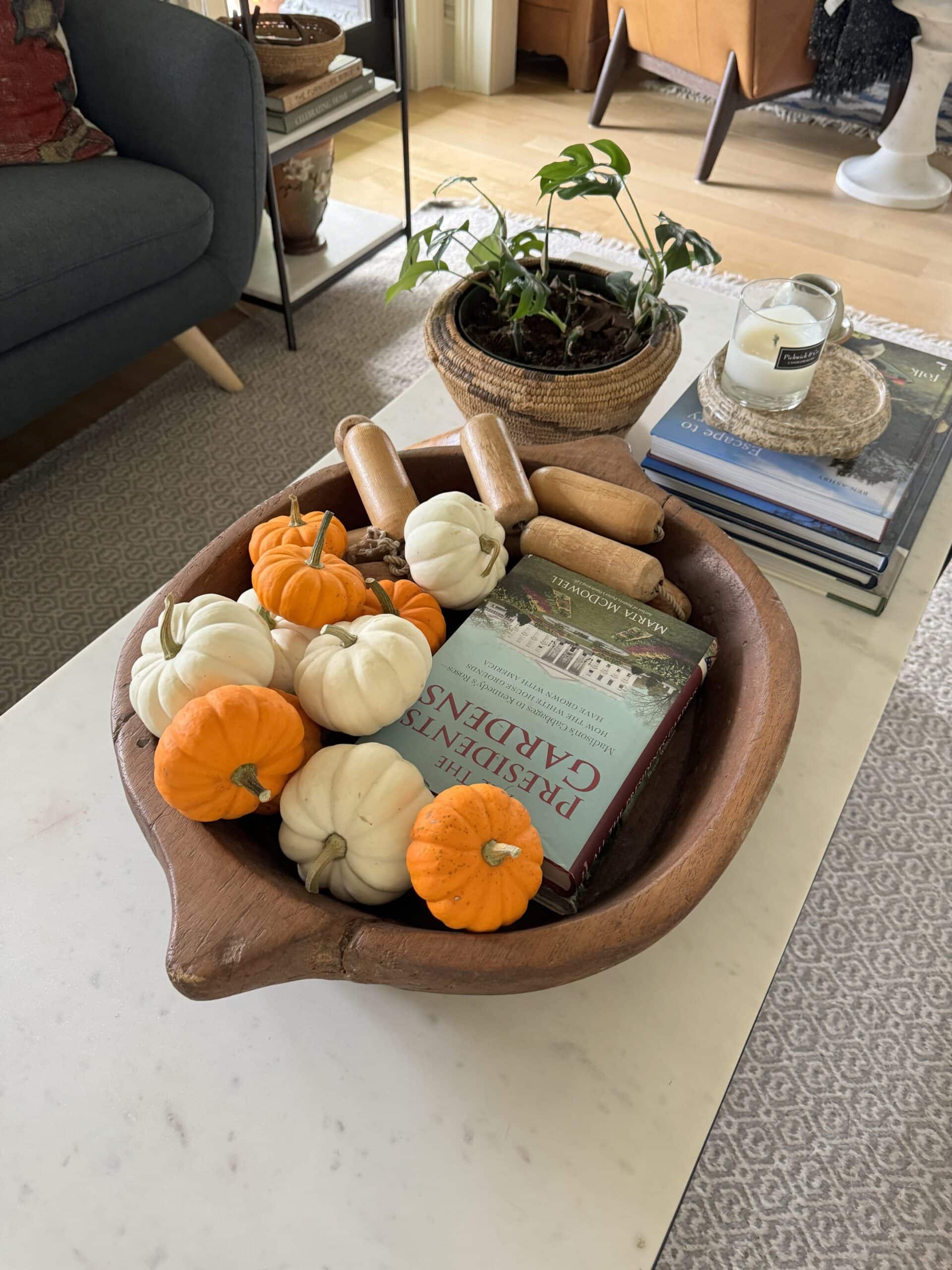 Coffee table with large wooden bowl filled with mini pumpkins with books, plant, and candle also on coffee table.
