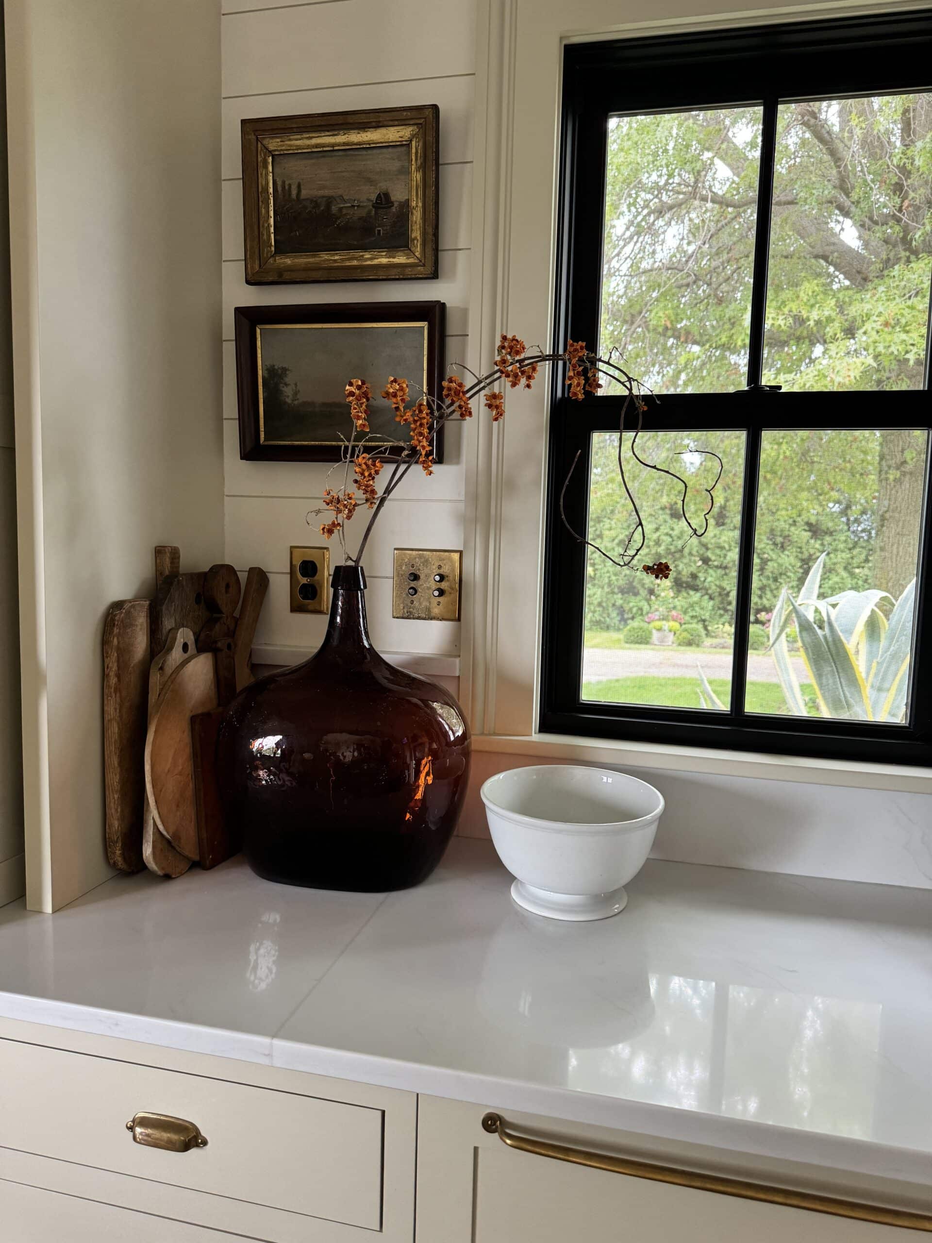 Kitchen countertop with large amber vessel with sprig of bittersweet in front of window.