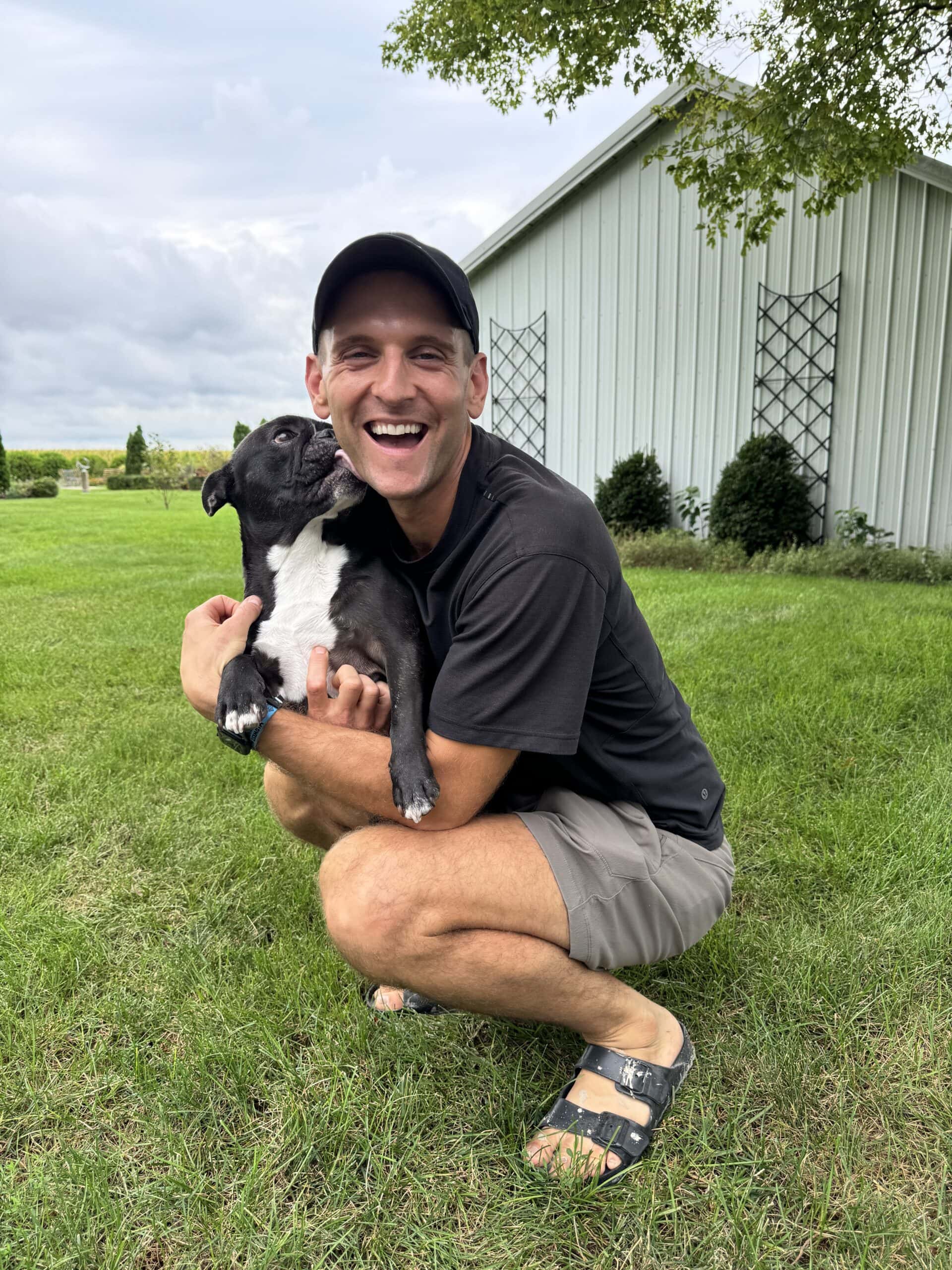 Kaleb crouching in grass with black French bulldog named Kip licking his face.
