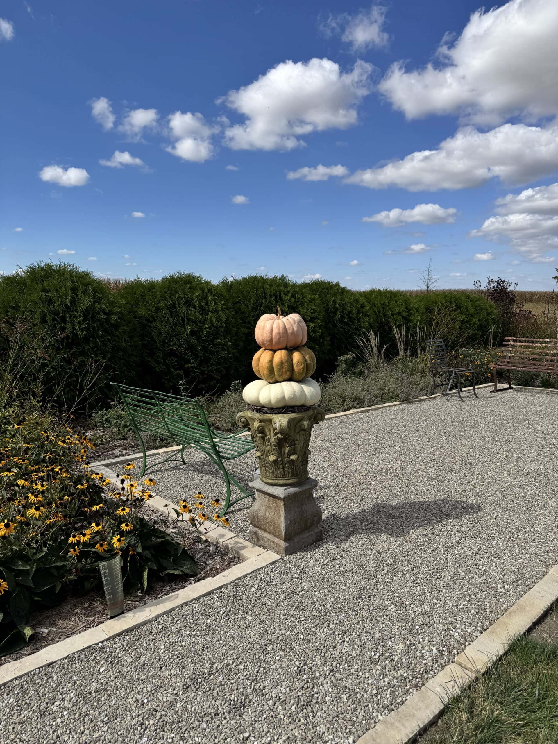 Gravel pathway with an urn with stacks of pumpkins on top with blue skies beyond.