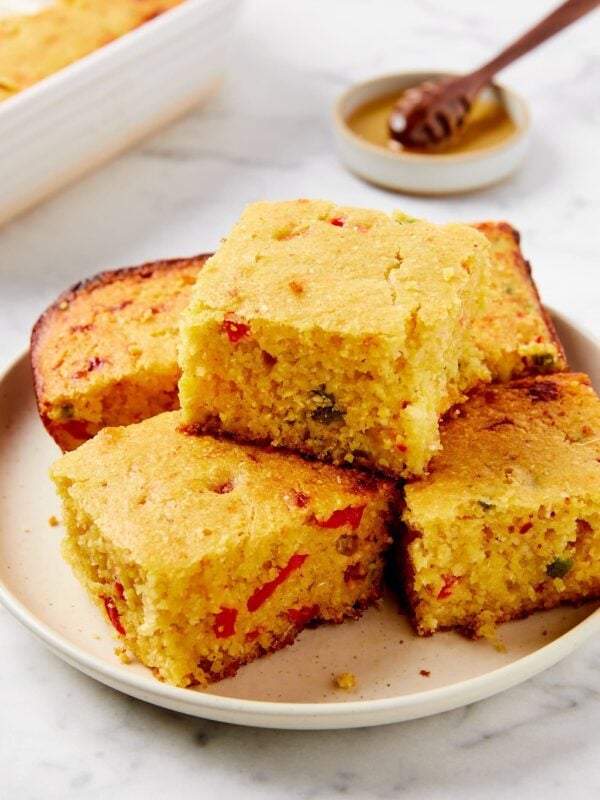 White plate with square pieces of jalapeño cornbread with a bit of honey in the background all on white marble surface.