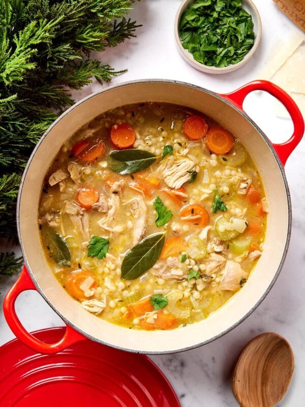 Large red Dutch oven on a white marble surface filled with turkey soup with bay leaves and pieces of carrots and turkey.