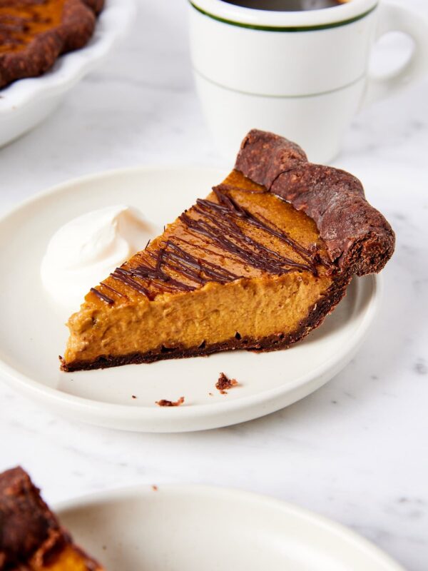 White plate with slice of pumpkin pie with chocolate crust drizzled with chocolate with whipped cream to the side.