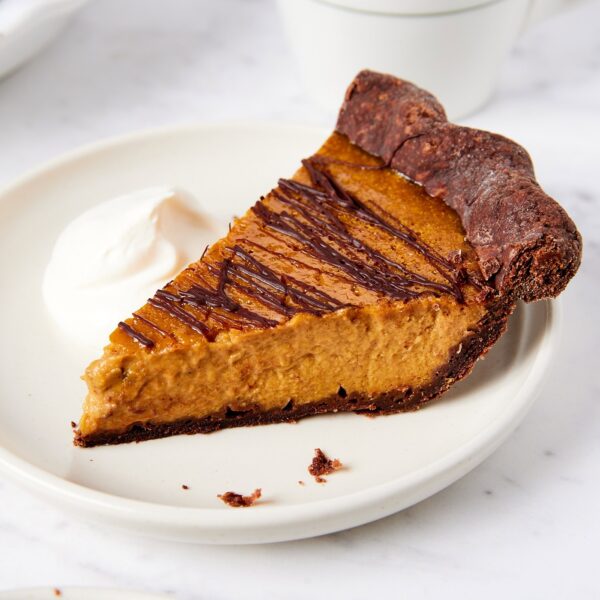 White plate with slice of pumpkin pie with drizzled chocolate on top with whipped cream to the side.