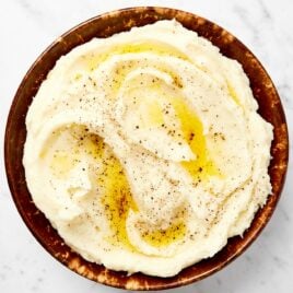 Brown bowl filled with creamy butter-covered mashed potatoes sitting on a marble surface.